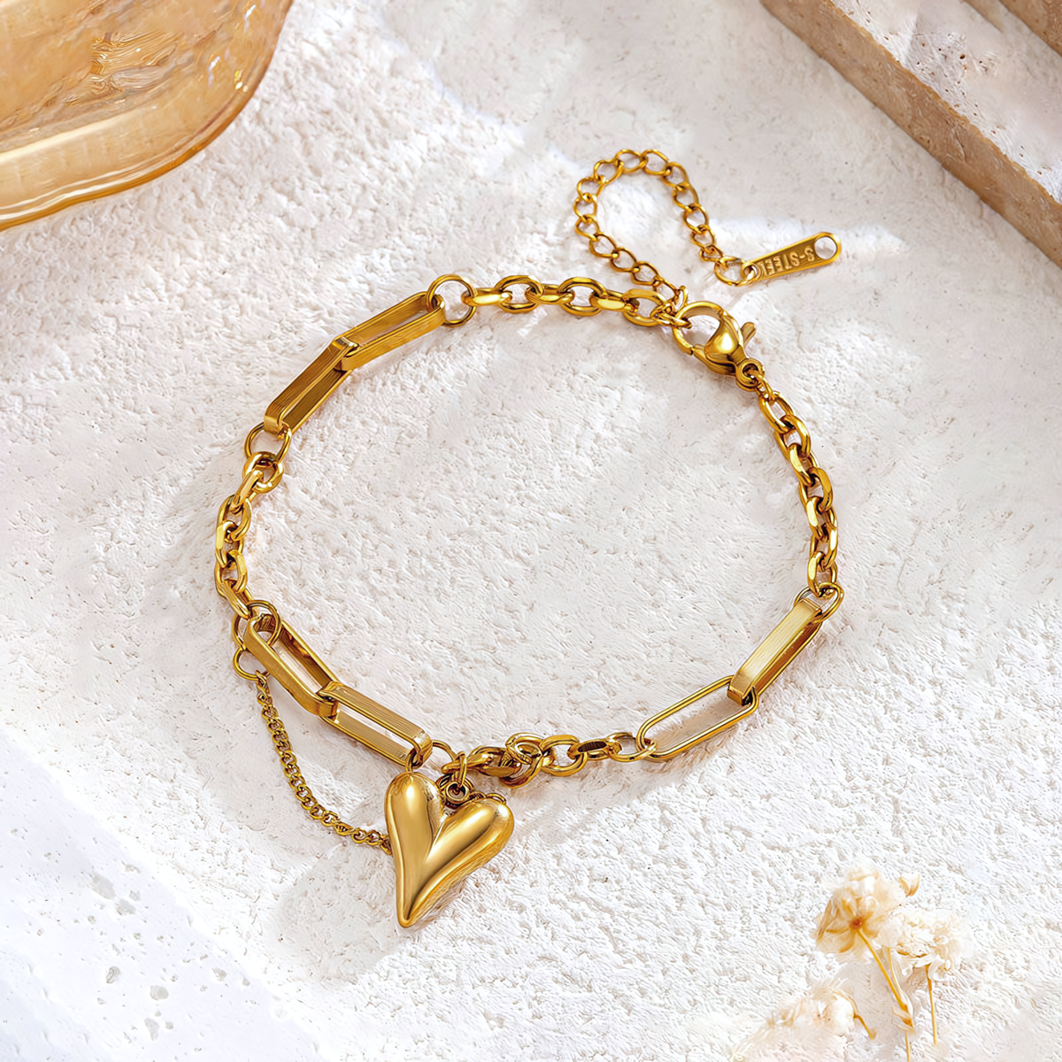 Flat lay of Amora gold heart charm chain bracelet on white textured stone with dried florals