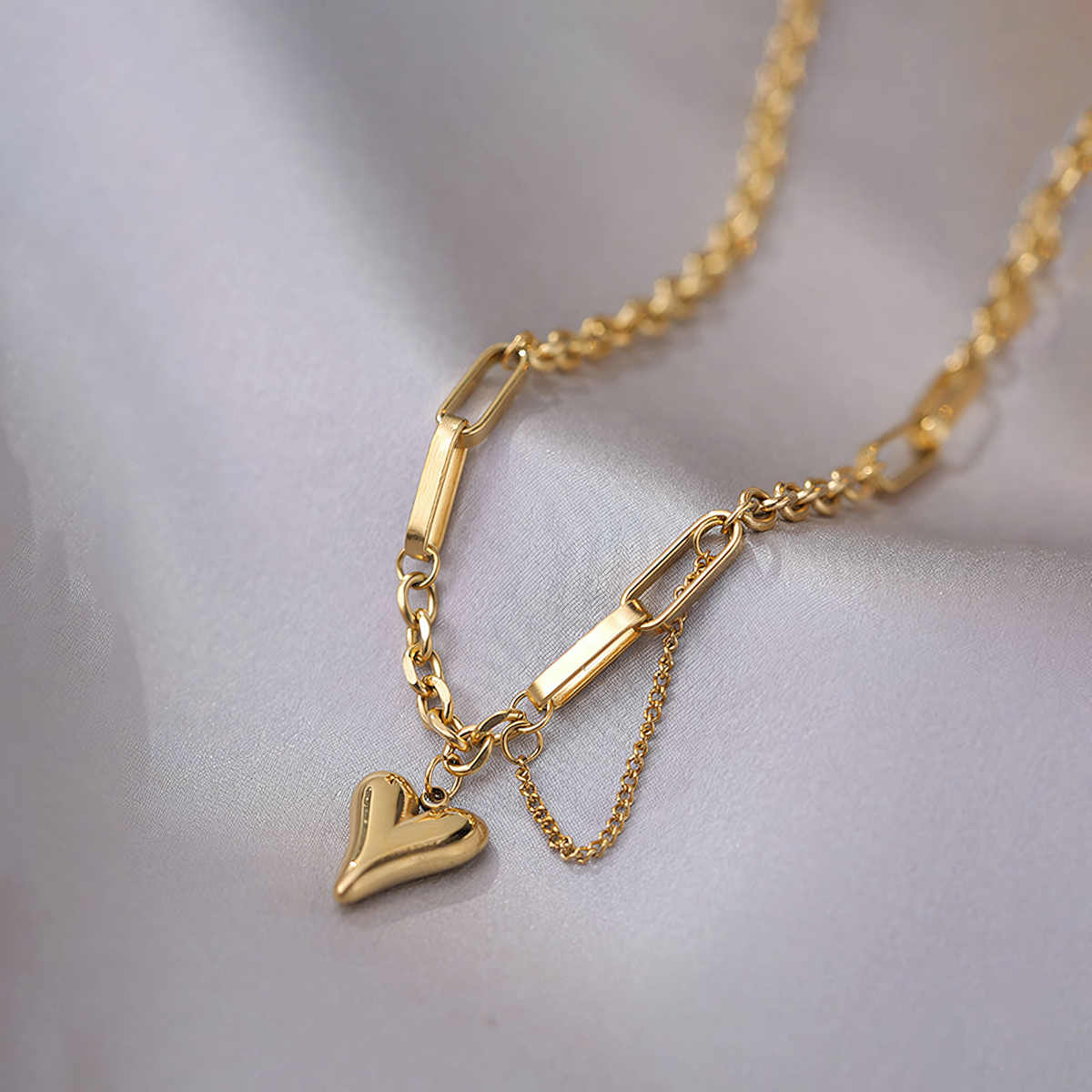 Close up of Amora gold paperclip chain bracelet with molten heart charm on grey satin fabric