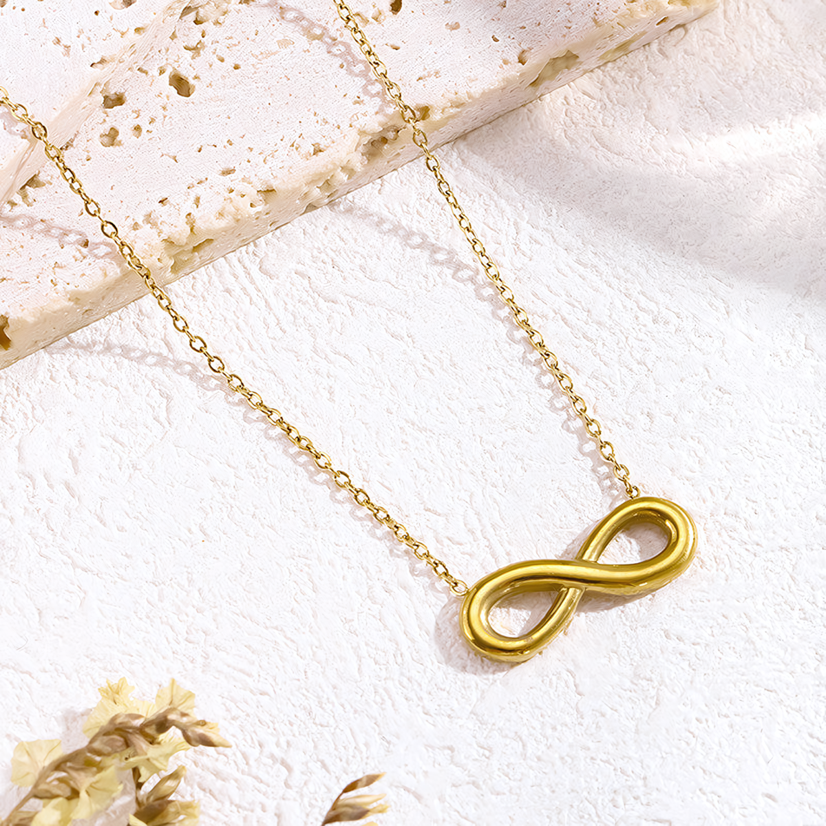 Aura 18K gold anti-tarnish infinity necklace resting on a textured white stone surface