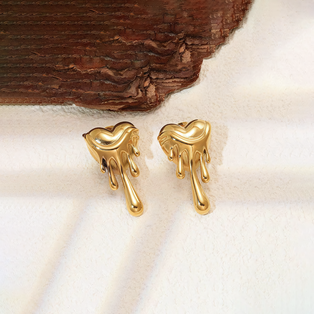 Celeste 18K gold plated stainless steel melting heart earrings resting on a white surface near dark wood