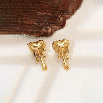 Celeste 18K gold plated stainless steel melting heart earrings resting on a white surface near dark wood