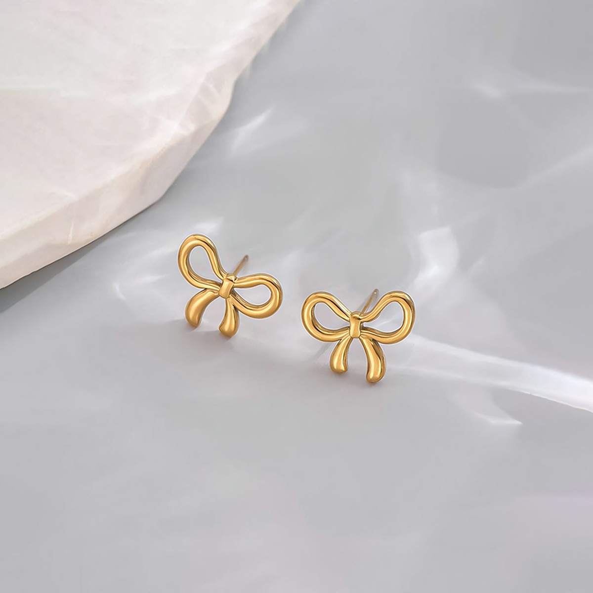 Clara 18K gold plated stainless steel bow earrings resting on a smooth grey surface